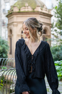 Blusa negra con escote en pico, textura de topos y lazos decorativos, estilo romántico y elegante