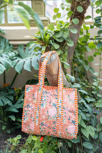 Bolsa tote de algodón con estampado floral en fondo color salmón y detalles de rayas naranjas, colgada sobre tronco entre hojas verdes