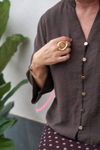 Detalle de blusa marrón chocolate con manga tres cuartos y botones metálicos dorados, acompañado de joyas doradas en primer plano

