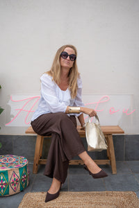 Pantalón fluido marrón con pinzas, look relajado sentada con blusa blanca, bolso dorado tipo saco y zapatos marrones
